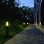 Lampa stojąca Manila H65, okrągła, czarna, umieszczona wzdłuż oświetlonej ścieżki. Widoczne źródło światła w górnej części lampy, otoczenie z zielenią i niską roślinnością, przy budynku. Dzień przekształca się w wieczór.