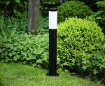 Okrągła lampa stojąca Manila H65 w kolorze czarnym, z białym kloszem u góry. Ustawiona na trawie, w otoczeniu zielonych krzewów. Wysoka, smukła konstrukcja, idealna do oświetlenia ogrodu lub ścieżek.