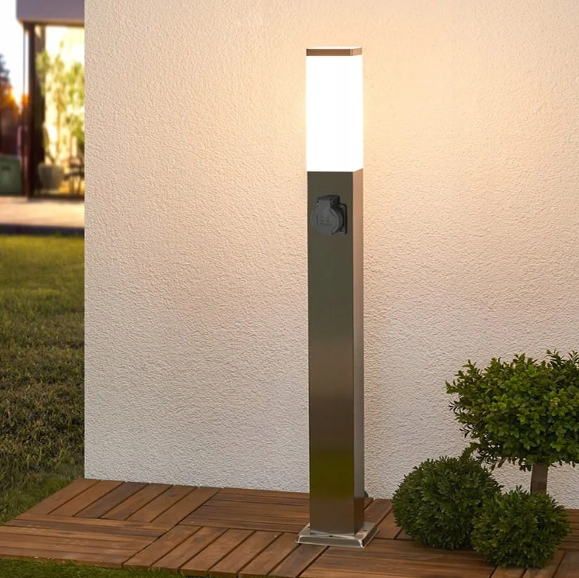 Lampa stojąca ogrodowa Havana H65 w kształcie kwadratu, wykonana z metalicznego srebra. Posiada gniazdo oraz mleczny klosz, emitujący światło, umieszczona na drewnianym podłożu, w otoczeniu niskich roślin.