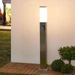 Lampa stojąca ogrodowa Havana H65 w kształcie kwadratu, wykonana z metalicznego srebra. Posiada gniazdo oraz mleczny klosz, emitujący światło, umieszczona na drewnianym podłożu, w otoczeniu niskich roślin.