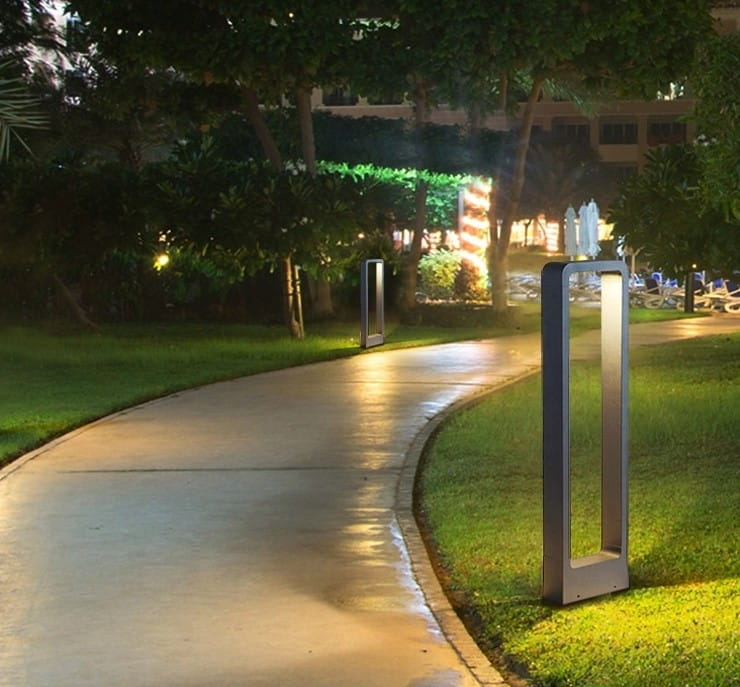 Lampa stojąca ogrodowa Rio LED H80 w kolorze antracytowym, w kształcie prostokątnego słupka, umieszczona w ogrodzie przy ścieżce. Oświetlenie emituje delikatne światło, otoczona zielenią i drzewami.