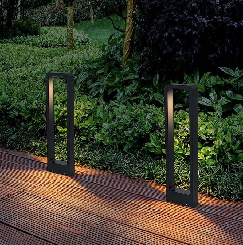Lampa stojąca ogrodowa Rio LED H60 w kolorze antracytowym, w formie prostokątnego słupka. Ujęcie z boku, na tle zieleni. Emituje ciepłe światło, umieszczona na drewnianym podłożu, co podkreśla nowoczesny design.