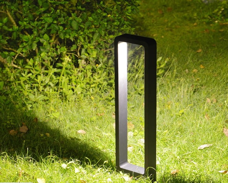 Antracytowa lampa stojąca ogrodowa Rio LED H60 w kształcie słupka, umieszczona na trawie. Produkt ma prostą, geometryczną formę z poziomym źródłem światła, które oświetla otoczenie. W tle widać zieleń krzewów.