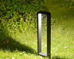 Antracytowa lampa stojąca ogrodowa Rio LED H60 w kształcie słupka, umieszczona na trawie. Produkt ma prostą, geometryczną formę z poziomym źródłem światła, które oświetla otoczenie. W tle widać zieleń krzewów.