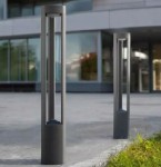 Lampa stojąca ogrodowa Mario H80 w antracytowym kolorze, o cylindrycznym kształcie, z szczelinami. Ujęcie z boku, na tle nowoczesnego budynku, z zielenią w dolnej części kadru.