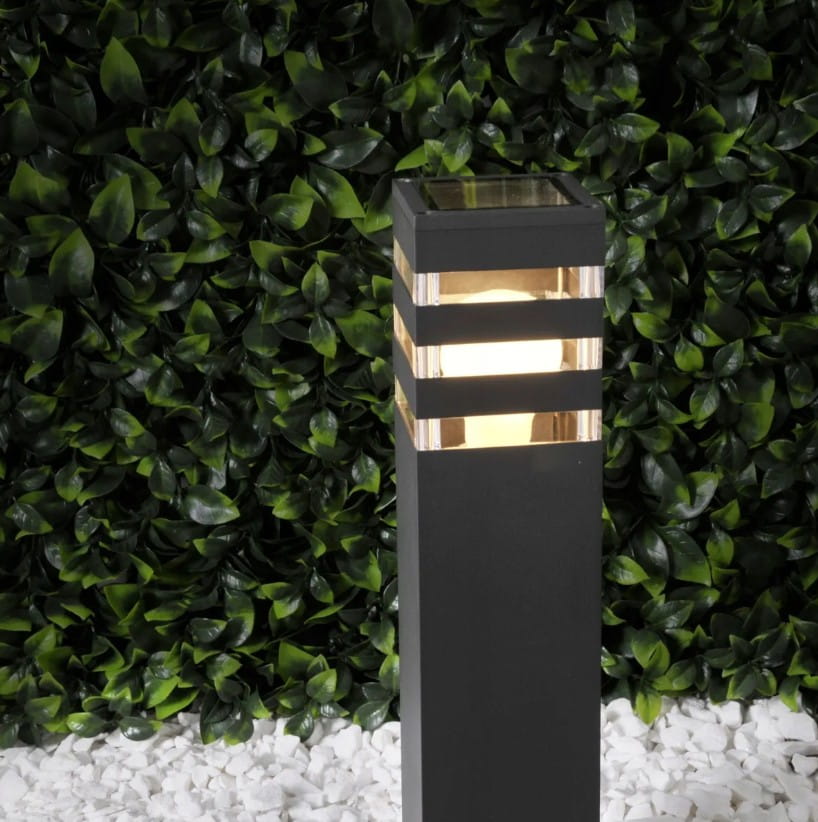 Lampa stojąca Malibu H50 w kształcie kwadratowego słupka, czarna, z przezroczystymi paskami u góry, 1xE27. Ujęcie z boku na tle zielonego żywopłotu. Widoczne oświetlenie wewnętrzne, które emituje ciepłe światło.