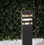 Lampa stojąca Malibu H50 w kształcie kwadratowego słupka, czarna, z przezroczystymi paskami u góry, 1xE27. Ujęcie z boku na tle zielonego żywopłotu. Widoczne oświetlenie wewnętrzne, które emituje ciepłe światło.