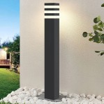 Lampa stojąca słupek ogrodowa Malibu H80 w kolorze czarnym, kwadratowa, z osłoną na żarówkę E27. Ujęcie z boku, lampa umieszczona na podłożu z kamieni, w tle widoczna zieleń i oświetlony mur.