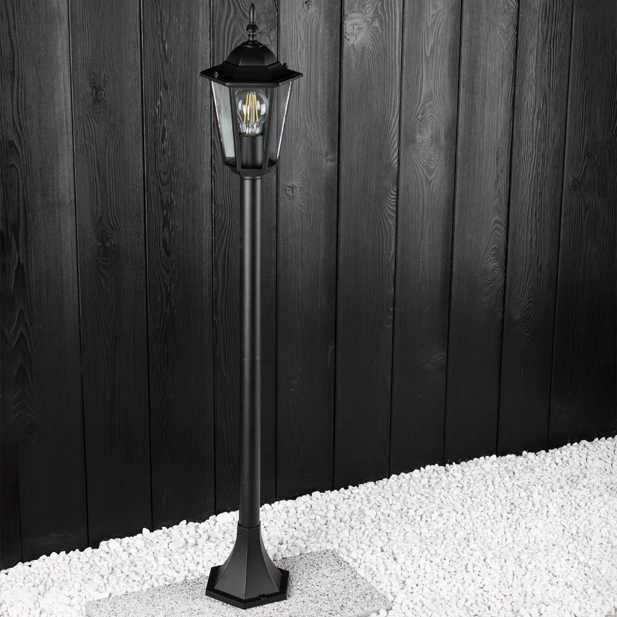 Lampa stojąca ogrodowa Victoria H100 w kolorze czarnym, 1-punktowa, z metalową konstrukcją i szklanym kloszem. Ujęcie z boku, na tle ciemnej drewnianej ściany, z białym żwirem u podstawy. Kształt cylindryczny, z ozdobnym daszkiem.