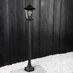 Lampa stojąca ogrodowa Victoria H100 w kolorze czarnym, 1-punktowa, z metalową konstrukcją i szklanym kloszem. Ujęcie z boku, na tle ciemnej drewnianej ściany, z białym żwirem u podstawy. Kształt cylindryczny, z ozdobnym daszkiem.