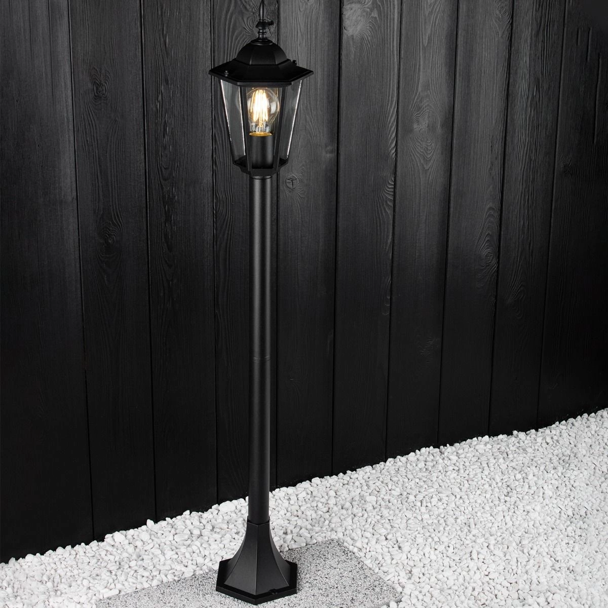 Lampa stojąca ogrodowa Victoria H100 w kolorze czarnym, z metalowym stelażem i kloszem, umieszczona na białych żwirach. Ujęcie z boku, pokazujące detal konstrukcji oraz źródło światła wewnątrz klosza. Tło z desek w ciemnym odcieniu.