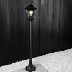 Lampa stojąca ogrodowa Victoria H100 w kolorze czarnym, z metalowym stelażem i kloszem, umieszczona na białych żwirach. Ujęcie z boku, pokazujące detal konstrukcji oraz źródło światła wewnątrz klosza. Tło z desek w ciemnym odcieniu.