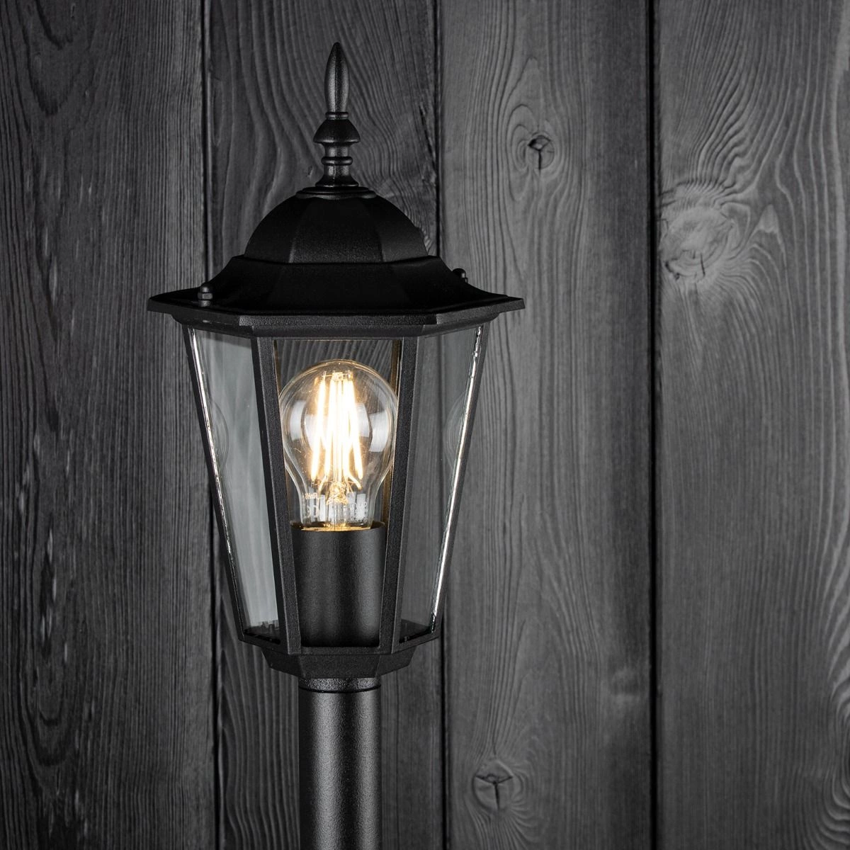 Czarna lampa stojąca ogrodowa Victoria H100 z metalową konstrukcją, szklanymi ściankami i ozdobnym daszkiem. Widoczna żarówka w środku, umieszczona na prostym słupku. Tło stanowi drewniana ściana w ciemnym odcieniu.