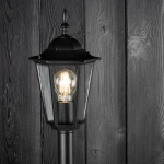 Czarna lampa stojąca ogrodowa Victoria H100 z metalową konstrukcją, szklanymi ściankami i ozdobnym daszkiem. Widoczna żarówka w środku, umieszczona na prostym słupku. Tło stanowi drewniana ściana w ciemnym odcieniu.