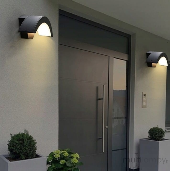 Kinkiet elewacyjny LED 9W w kolorze czarnym, zamontowany na zewnętrznej ścianie budynku. Oświetlenie w formie łuku z białym kloszem, umieszczone po obu stronach drzwi wejściowych. Roślinność w donicach w tle.