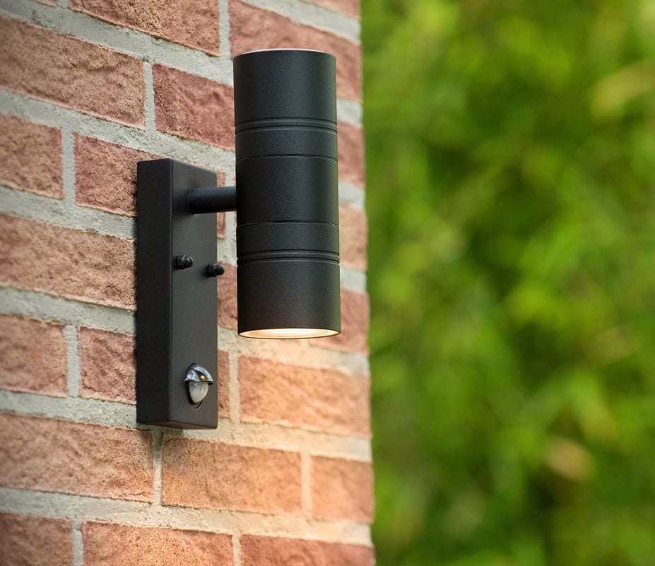 Kinkiet elewacyjny Arne w kolorze czarnym, 2-punktowy, zamontowany na ceglanej ścianie. Okrągła lampa z widocznym czujnikiem ruchu, skierowana w dół. Czyste linie i minimalistyczny design, idealny do oświetlenia zewnętrznego.