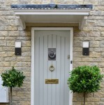 Grafitowy kinkiet elewacyjny Clark z czujnikiem ruchu, zamontowany po obu stronach drzwi wejściowych. Produkt o prostokątnym kształcie, z matowym wykończeniem i białym kloszem, umieszczony na tle jasnej, kamiennej elewacji.