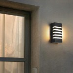 Koga kinkiet elewacyjny 1xE27 w kolorze czarnym, zamontowany na ścianie, z poziomymi, szczelinowymi dekoracjami, emitującym ciepłe światło. Obok okna z firanką, tło w neutralnym odcieniu.