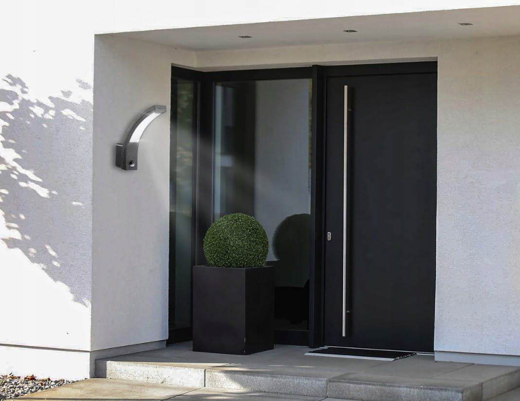 Diron LED kinkiet elewacyjny w kolorze czarnym z czujnikiem ruchu, zamontowany na ścianie obok drzwi. Kinkiet ma zaokrąglony kształt, emitując światło w kierunku podłogi. W tle widoczna donica z roślinnością oraz drzwi w kolorze czarnym.