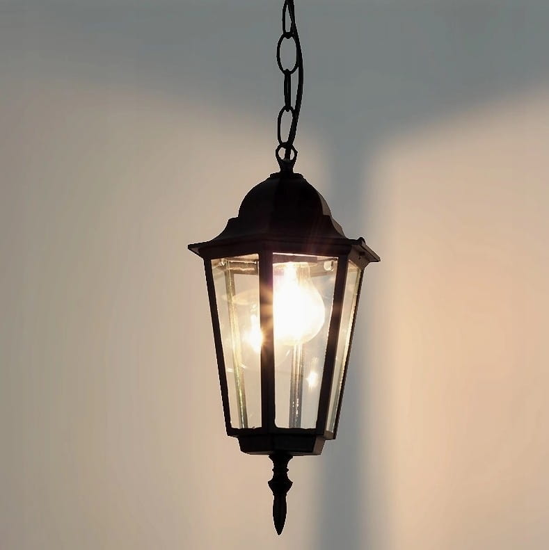 Czarna lampa wisząca ogrodowa Victoria, 1-punktowa, o kształcie latarni. Wykonana z metalu z przezroczystymi ściankami, zawieszona na łańcuszku. Wewnątrz widoczna żarówka, emitująca światło. Tło jasne, co podkreśla detal lampy.