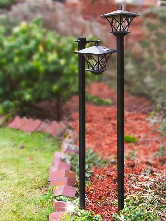 Lampa solarna ogrodowa z czujnikiem zmierzchu LED w kolorze czarnym, stojąca na dwóch metalowych słupkach. Ma osłonę w kształcie latarni z przezroczystymi elementami. Ujęcie z boku, w ogrodzie, na tle zieleni i brązowego podłoża.