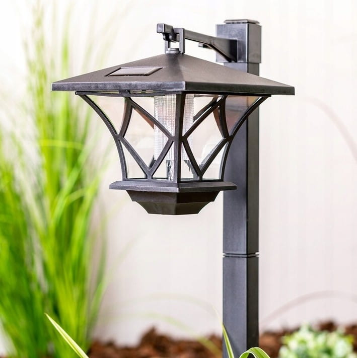 Lampa solarna ogrodowa z czujnikiem zmierzchu LED, czarna, z metalowym korpusem i przezroczystymi panelami. Ujęcie z boku, pokazujące szczegóły konstrukcji oraz otoczenie z roślinami, na tle jasnej ściany.