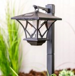 Lampa solarna ogrodowa z czujnikiem zmierzchu LED, czarna, z metalowym korpusem i przezroczystymi panelami. Ujęcie z boku, pokazujące szczegóły konstrukcji oraz otoczenie z roślinami, na tle jasnej ściany.