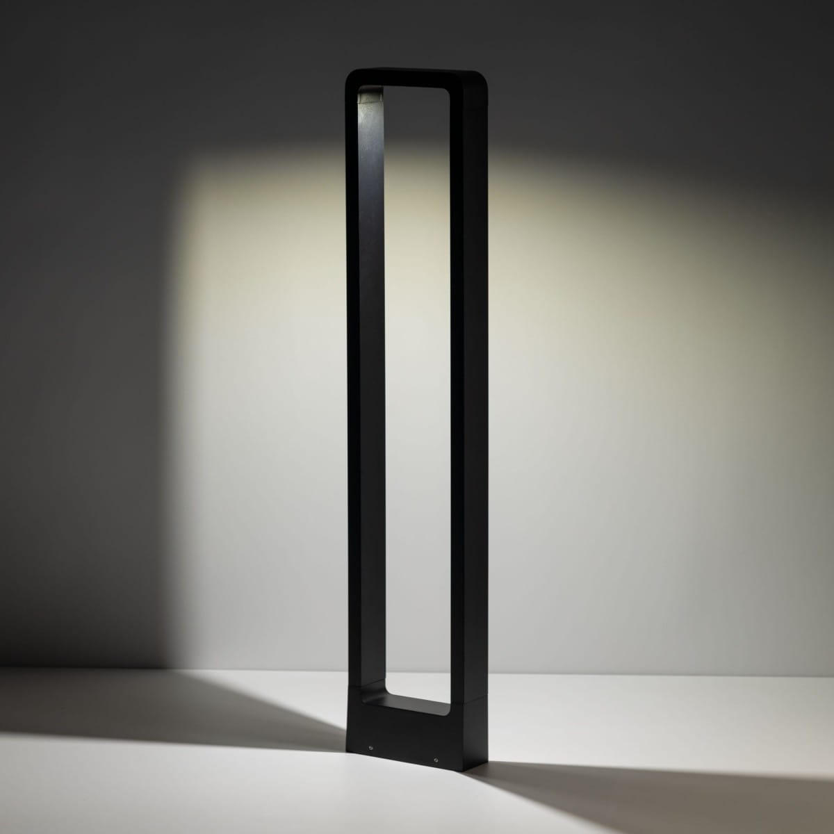 Czarna lampa stojąca ogrodowa Rio LED H80 w formie słupka, o prostokątnym kształcie i minimalistycznym designie. Ujęcie z boku, ukazujące smukłą sylwetkę i emitowane światło w kierunku ściany.