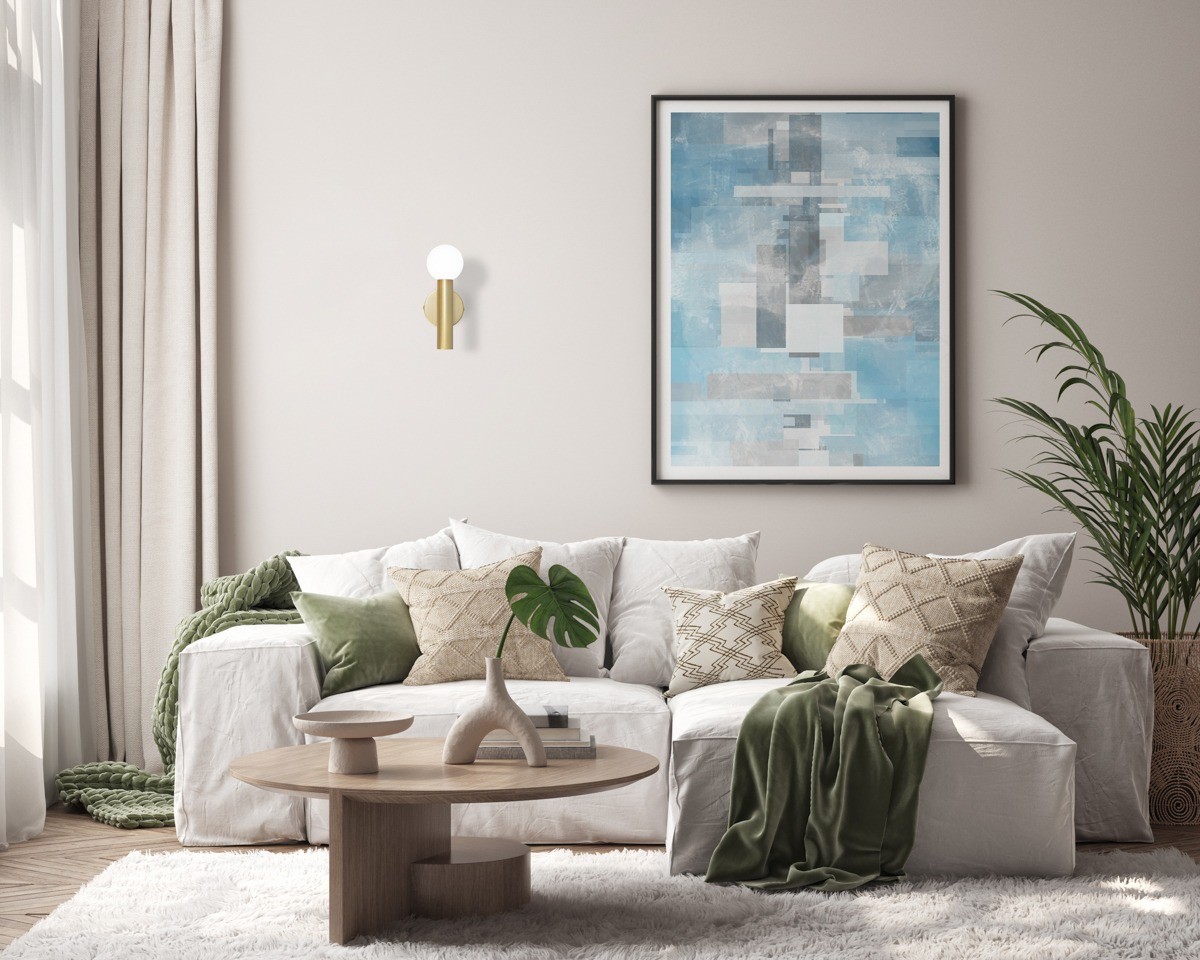 Złoty kinkiet Filano 1xG9, zamontowany na jasnej ścianie, z okrągłą, białą kloszem. W tle widoczny nowoczesny obraz w odcieniach niebieskiego oraz aranżacja salonu z białą sofą, zielonymi poduszkami i dekoracyjnymi roślinami.