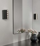 Kinkiet LED w kolorze czarnym o mocy 10W i temperaturze barwowej 4000K. Zainstalowany na ścianie obok lustra, z pięcioma źródłami światła. W tle widać czarny wazon z różami na ciemnym blacie.