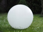 Lampa solarna ogrodowa Ballo w kształcie dużej, białej kuli o średnicy 30 cm i wysokości 65 cm, umieszczona na trawie, otoczona kwiatami. Widać jej gładką, błyszczącą powierzchnię.