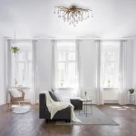 Lampa sufitowa Icicle w postarzanym mosiądzu, z 4 źródłami G9, zawieszona na białym tle. Posiada dekoracyjne ramiona w kształcie gałęzi, z przeszklonymi kulkami. Wnętrze w jasnych barwach, z szarym narożnikiem i roślinami w tle.