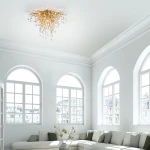 Lampa sufitowa Icicle w postarzanym mosiądzu z dziewięcioma gniazdami G9, o organicznej formie przypominającej gałęzie, z dekoracyjnymi kryształowymi kroplami, montowana na suficie w jasnym wnętrzu z dużymi oknami.