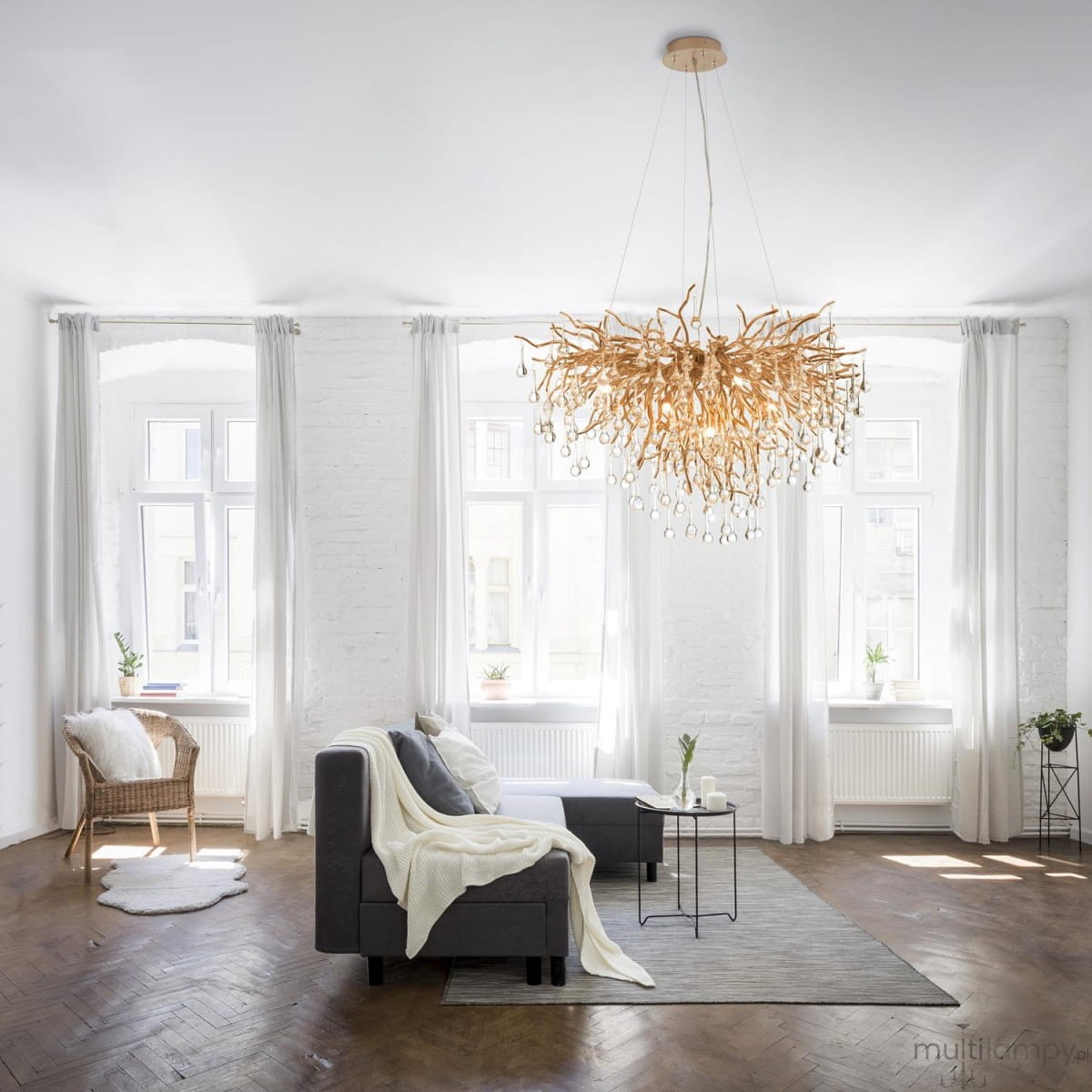 Lampa wisząca Icicle w postarzanym mosiądzu, z 10 gniazdami G9, o organicznym kształcie przypominającym gałęzie, zawieszona w jasnym wnętrzu z dużymi oknami. W tle widoczne meble i dekoracje w stonowanej kolorystyce.