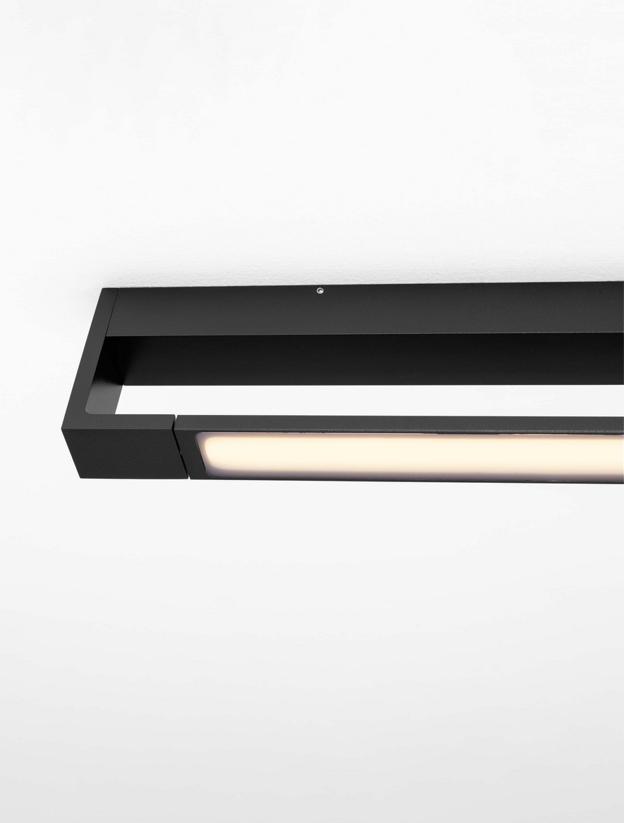 Kinkiet Justo 1xLED 12W w kolorze czarnym, o prostokątnym kształcie, zamontowany na białym tle. Posiada matową obudowę z LED-em wzdłużnej linii, emitującą ciepłe światło. Widoczny detal montażowy.