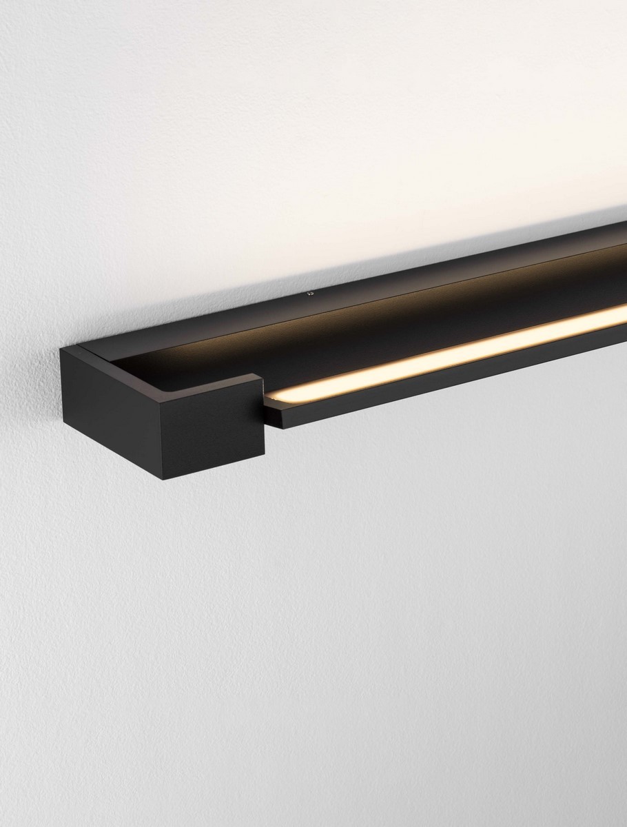 Kinkiet Justo 1xLED 17W w kolorze czarnym, zamontowany na ścianie. Prosty, prostokątny kształt z wąskim źródłem światła LED, emitującym ciepłe światło. Tło jasne, co podkreśla minimalistyczny design lampy.