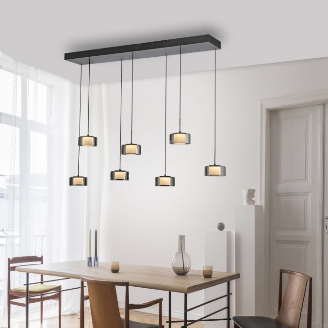 Lampa wisząca Rika z 7 źródłami światła LED, 56W, w czarnym dymionym kolorze. Posiada prostokątne klosze, zawieszone na cienkich linkach, umieszczona nad drewnianym stołem w jasnym wnętrzu. Tło białe z dużymi oknami.