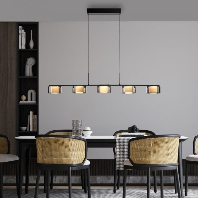 Lampa wisząca Rika z pięcioma źródłami LED, czarna z dymionym kloszem, zawieszona nad stołem. Klosze mają lekko złotawy odcień, a lampa zwisa z prostokątnej podstawy. Otoczenie to nowoczesna jadalnia z krzesłami ratanowymi.
