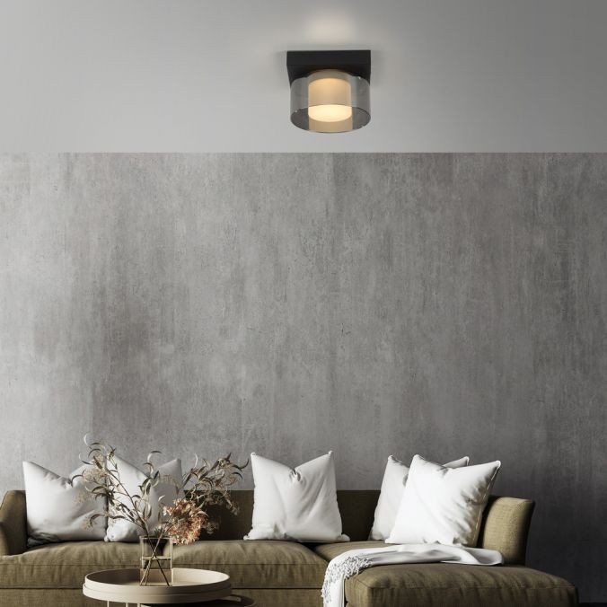 Lampa sufitowa LED Rika 4,5W w czarnym dymionym kolorze, montowana na suficie. Okrągła konstrukcja z przezroczystym kloszem, emitująca ciepłe światło. Widoczna w aranżacji salonu z kanapą i poduszkami.