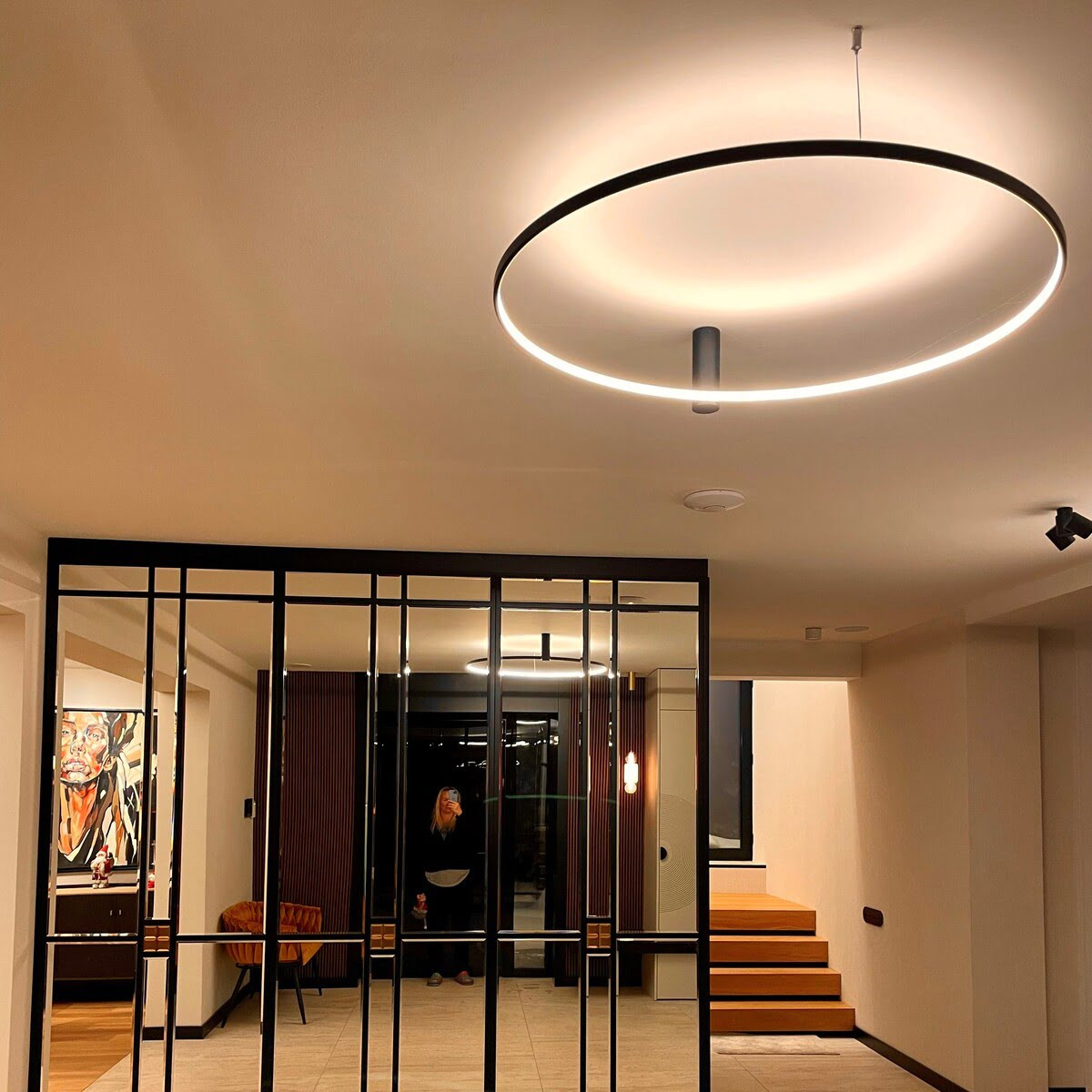 Czarna lampa sufitowa LED Ring D90 o mocy 50W, w kształcie okręgu, zawieszona na suficie. Emituje ciepłe światło, widoczna w nowoczesnym wnętrzu z drewnianymi schodami i przeszklonymi ścianami.