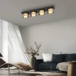Lampa sufitowa Rika 4xLED 26W w czarnym dymionym wykończeniu. Produkt umieszczony na suficie, z czterema okrągłymi kloszami, emitującymi ciepłe światło. Otoczenie obejmuje jasny salon z dużym oknem i ciemnoszarym sofą.