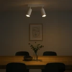 Lampa sufitowa Mica w kolorze białym i chromowanym, z dwoma punktami świetlnymi GU10, zamontowana na prostokątnej podstawie. Ujęcie z góry, oświetlająca stół z drewnianym blatem, w otoczeniu czarnych krzeseł i dekoracji.