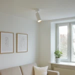 Mica lampa sufitowa (spot) 1xGU10 w kolorze białym i chromowanym, zamontowana na suficie. Lampa skierowana w dół, w minimalistycznej aranżacji z jasnymi ścianami, obok dwa obrazy w ramach i roślina w doniczce na parapecie.