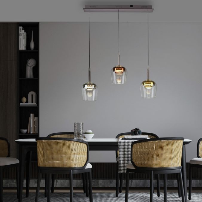 Lampa wisząca Q-kon z pilotem, 3 klosze w różnych kolorach, zawieszone na cienkich linkach. Wyróżniają się szklanym wykończeniem i metalowymi detalami. Umieszczona nad stołem w nowoczesnej aranżacji, na tle jasnej ściany.