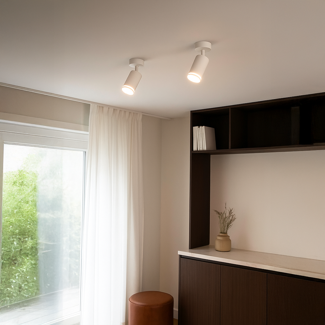 Biała lampa sufitowa Pipe Ring z jedną oprawą GU10, zamontowana pod kątem na suficie. W tle widoczna półka z książkami oraz jasne zasłony przy oknie. Lampa o prostym, cylindrycznym kształcie, zapewniająca skupione światło.