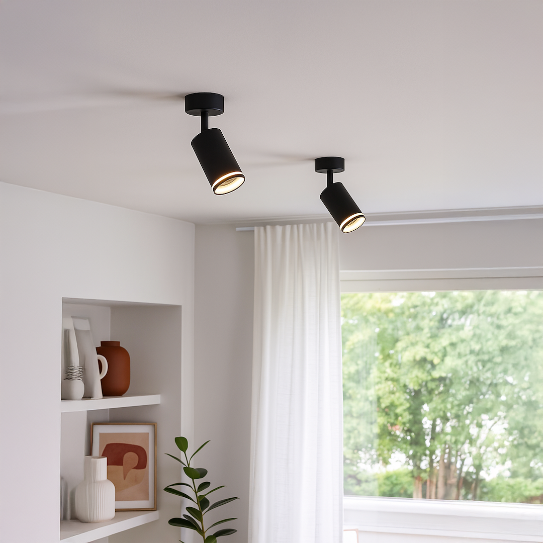 Czarna lampa sufitowa Pipe Ring z jednym spotem GU10, zamontowana na suficie, z lekko nachyloną głowicą. Otoczenie ze ścianą w jasnym kolorze, zasłonami i rośliną w doniczce w tle.