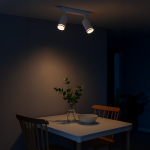 Lampa sufitowa Pipe Ring z dwoma spotami GU10 w białym kolorze, zamontowana na suficie. Ujęcie z boku, w tle stół z białym naczyniem i wazonem z rośliną. Oświetlenie tworzy przytulną atmosferę w pomieszczeniu.