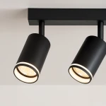 Lampa sufitowa Pipe Ring z dwoma reflektorami GU10 w kolorze czarnym, zamontowana na prostokątnej podstawie. Reflektory mają cylindryczny kształt i są ustawione pod różnymi kątami, emitując ciepłe światło. Tło jasne, minimalistyczne.