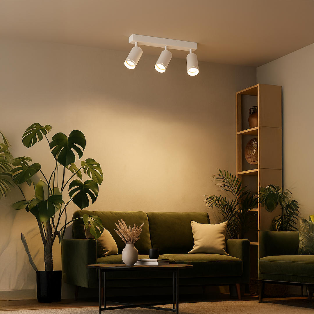 Lampa sufitowa Pipe Ring z trzema reflektorami GU10 w kolorze białym, zamontowana na prostokątnej podstawie. Reflektory są ustawione w linii, skierowane w różnych kierunkach. Lampa znajduje się w jasnym wnętrzu z zieloną sofą i roślinami.