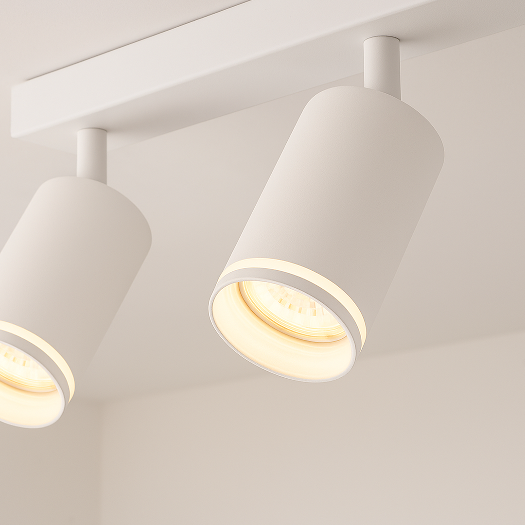 Lampa sufitowa Pipe Ring z trzema reflektorami GU10 w kolorze białym. Cylindryczne klosze z matowej powierzchni, zamontowane na prostokątnej podstawie. Widoczne ciepłe światło emitowane przez żarówki. Ujęcie z bliska, z góry, ukazujące detale designu.